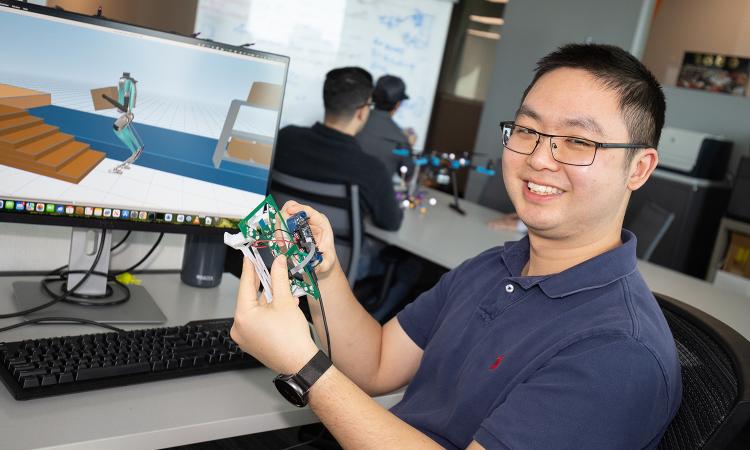 a student holds up a circuitboard in front of a computer screen with robotics software