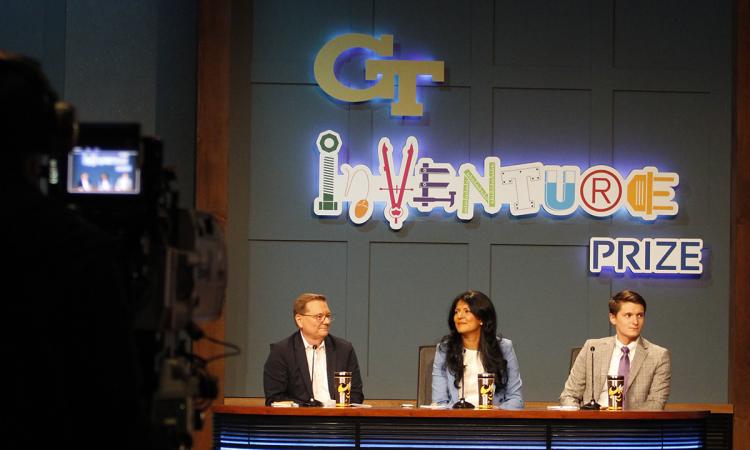 A TV camera in shadow in the foreground points at a table with three judges and a large sign behind them with the Georgia Tech and InVenture Prize logos.