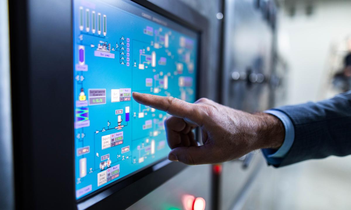 A finger nearly touching a control screen on an industrial system. The screen shows a visualization of the various components of the system.